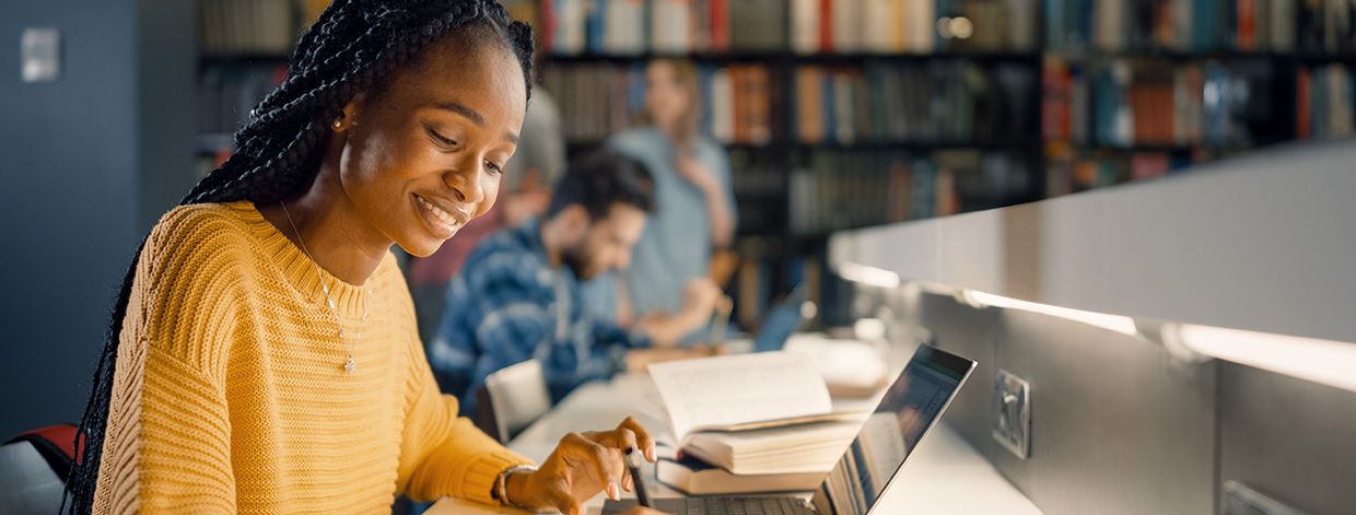 Biblioteca universitaria: una ragazza nera dotata usa un laptop e prende appunti