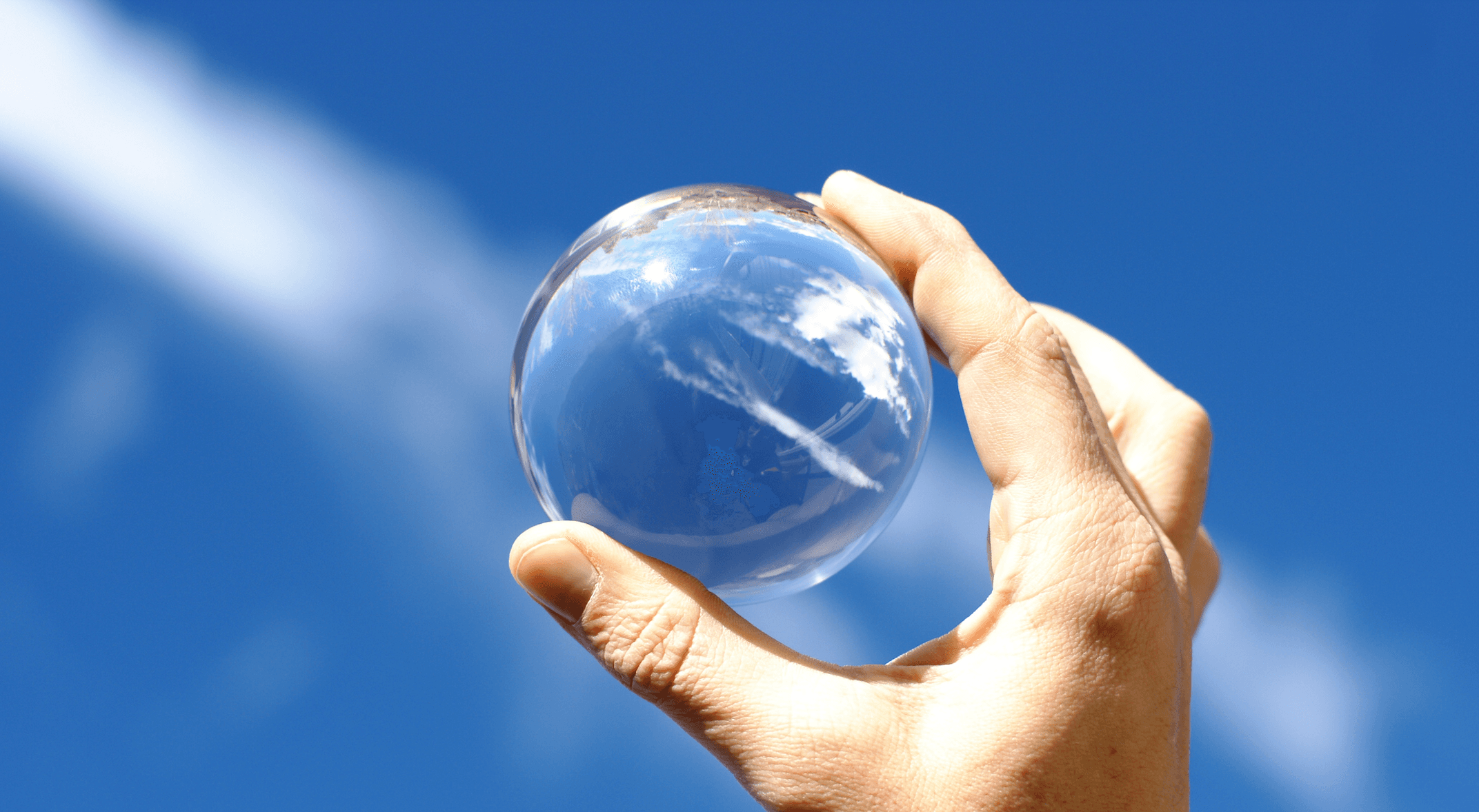 A hand holds a crystal ball aloft against a blue sky with wispy clouds.
