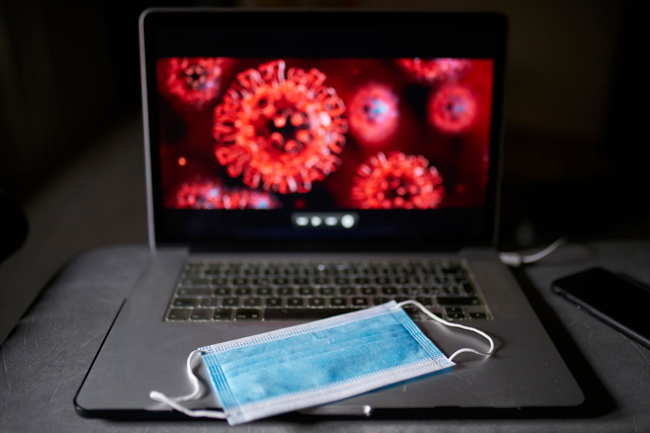 In uno spazio buio, una mascherina blu è posata sulla tastiera di un laptop, con immagini microscopiche del virus COVID rosso e appuntito che risplendono dallo schermo.
