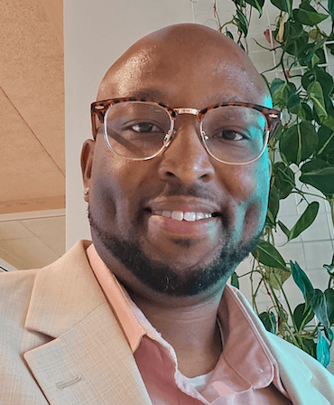 Photo of a bald man with a dark mustache and beard wearing glasses and a tan blazer.
