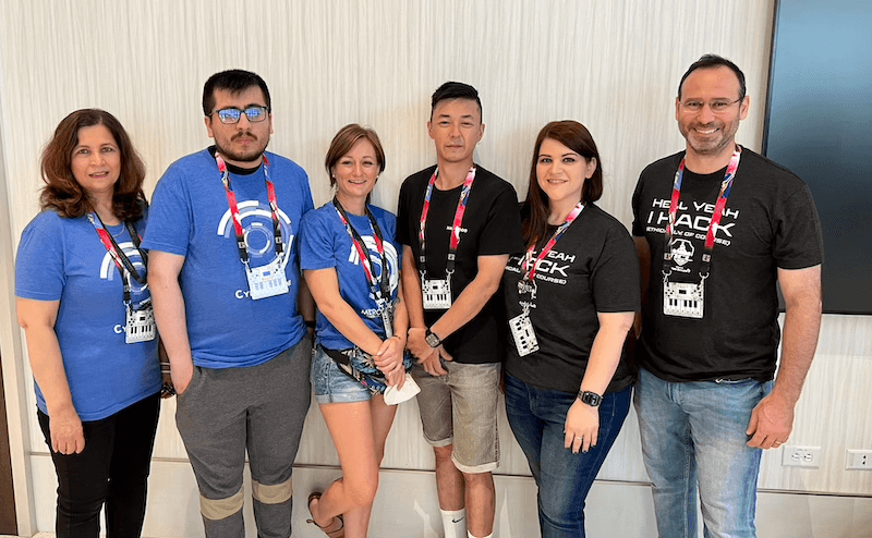 Photo of 6 people wearing matching t-shirts and convention lanyards posing for a photo in a convention hall.
