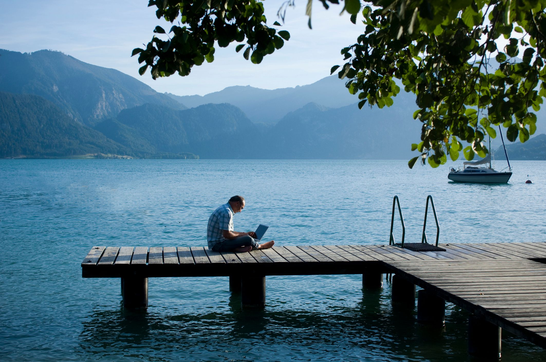 Foto di un uomo seduto a gambe incrociate su un molo vicino a un lago con un laptop in grembo.