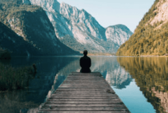 Una persona seduta su un molo affacciato su un lago di montagna e su un paesaggio boscoso