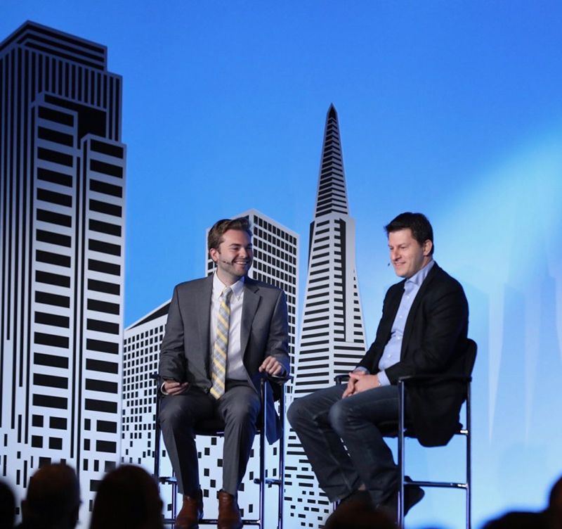 Due uomini intenti a conversare su un palco, con lo skyline di San Francisco sullo sfondo