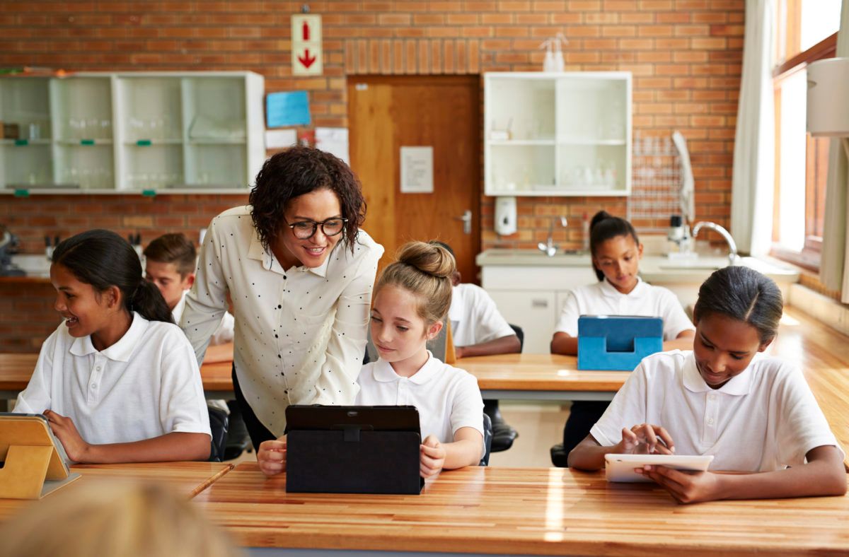 Insegnante che assiste gli studenti ai banchi con un tablet e dei fogli in classe