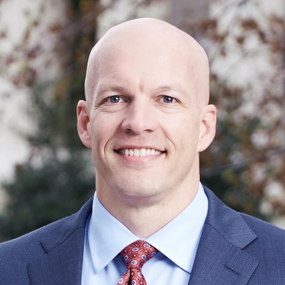 Professional headshot of a man in a dark suit and red patterned tie